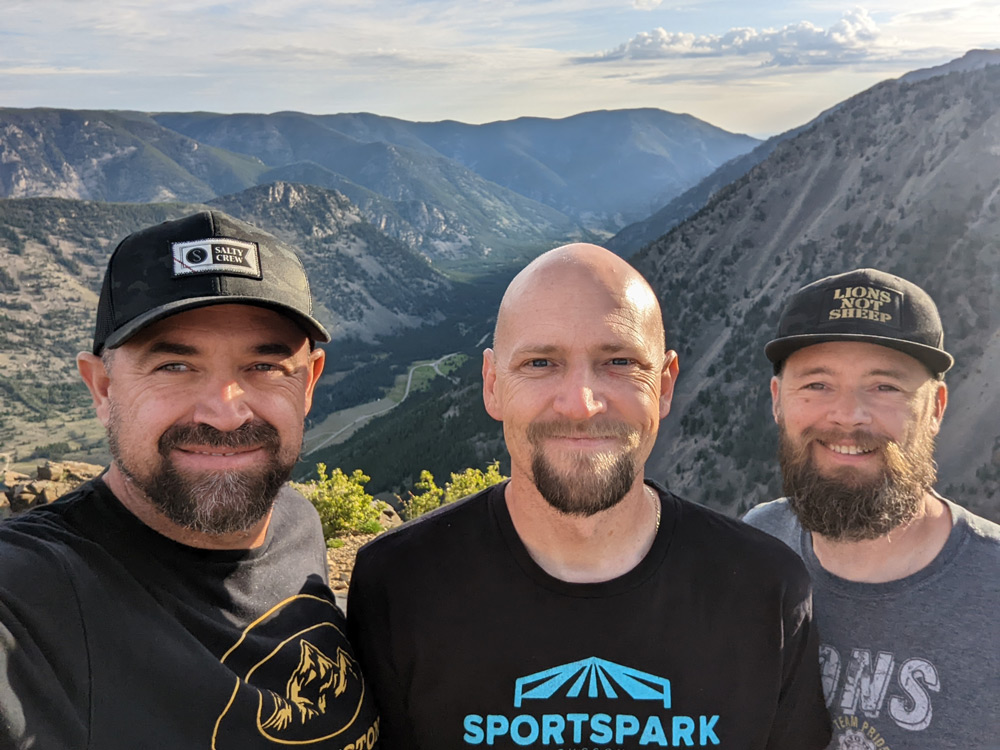 Three smiling men standing together outdoors against a scenic mountain backdrop, representing the Beloved remodeling team. They are dressed casually in branded apparel, showcasing their professional identity and connection to craftsmanship and home improvement.